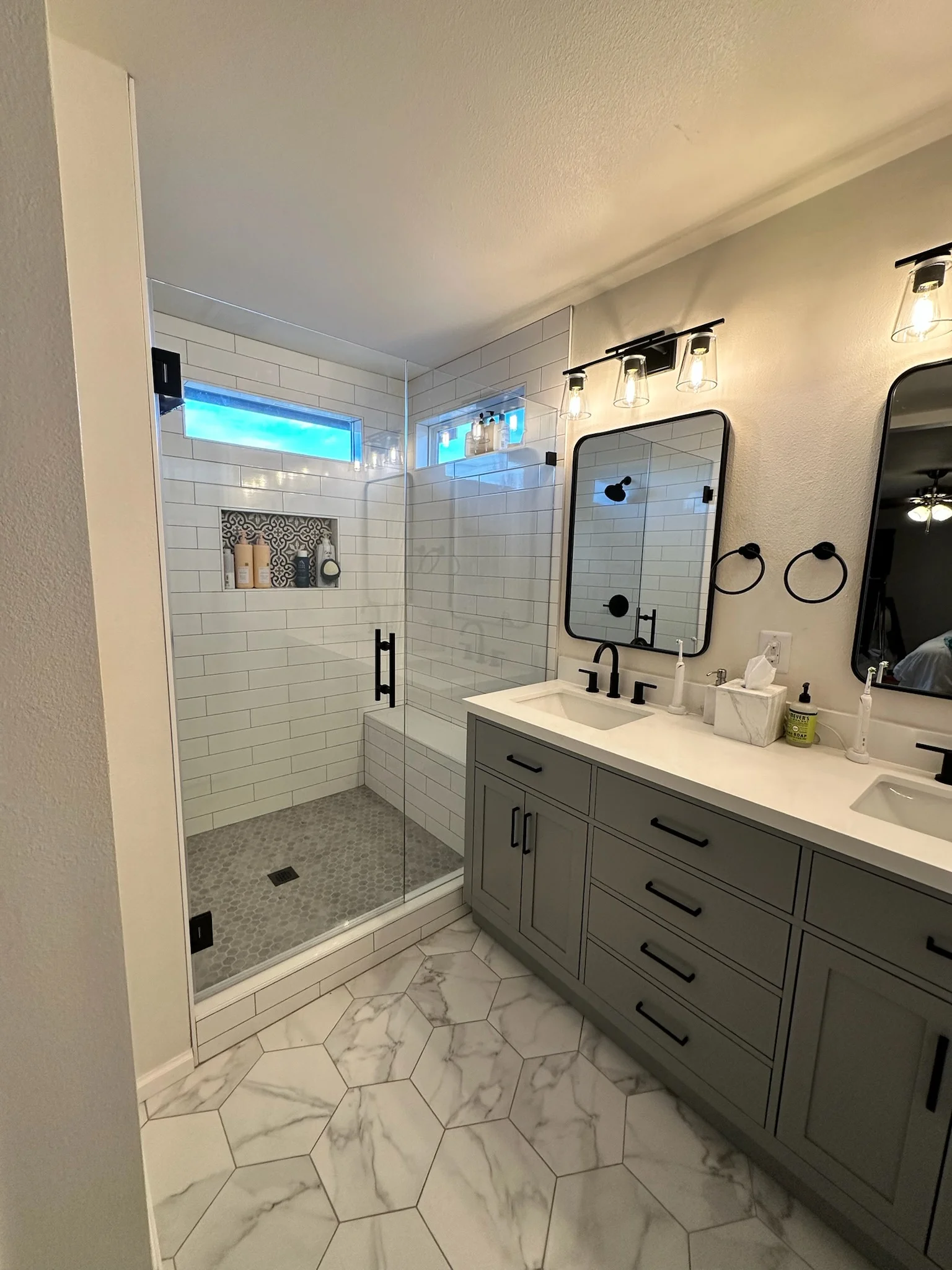 Modern bathroom with glass shower, subway tile, marble hexagon flooring, gray vanity, and dual sinks