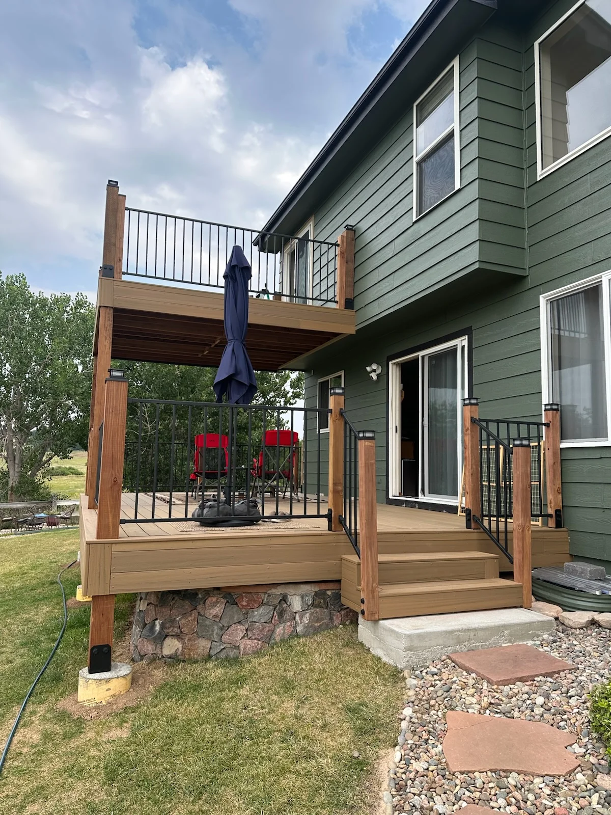 Two-level composite deck with cedar posts, metal railings, and stone foundation attached to green-sided home